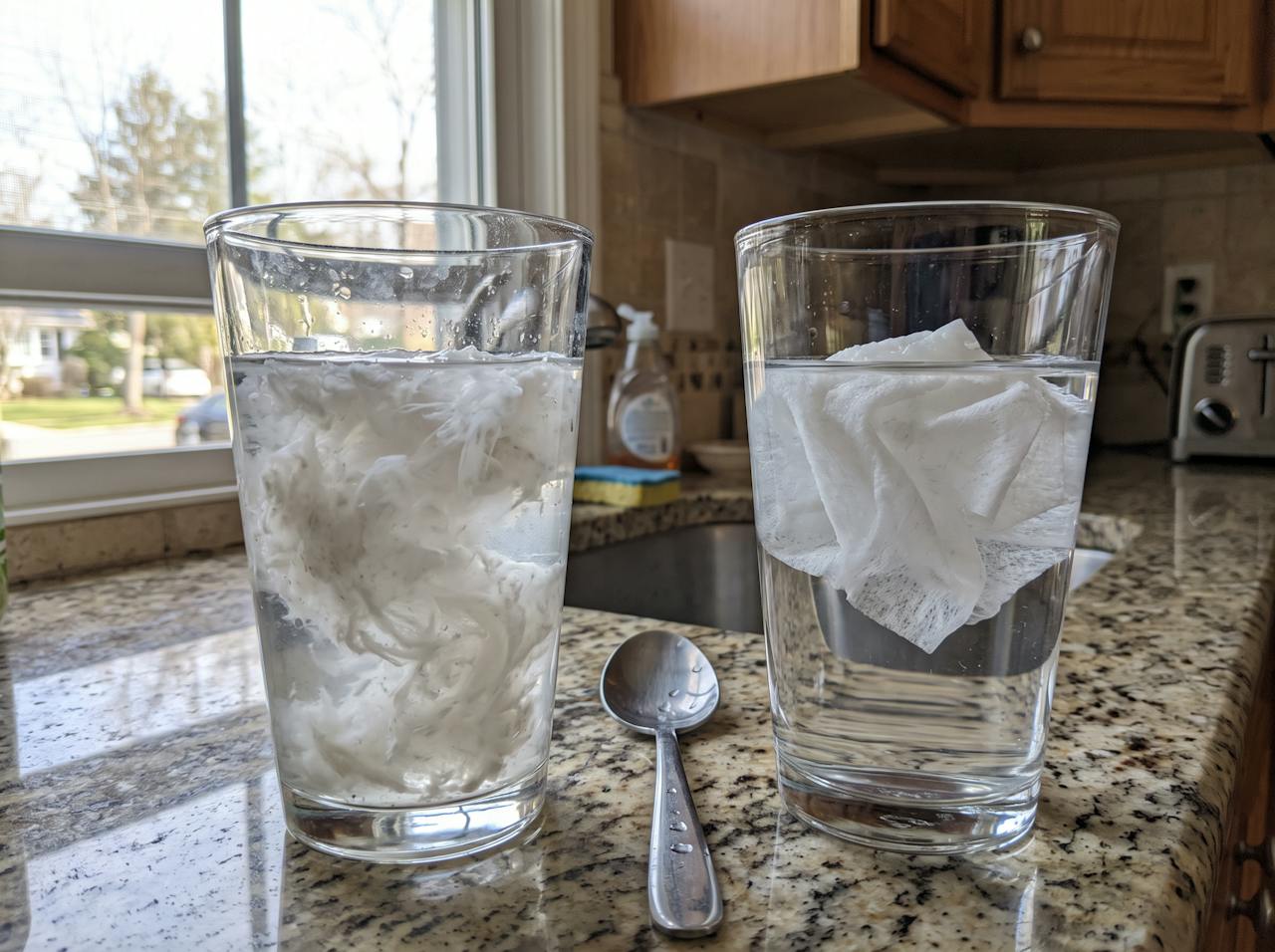 Toilet paper broken apart in one glass of water next to a flushable wipe still whole in another glass