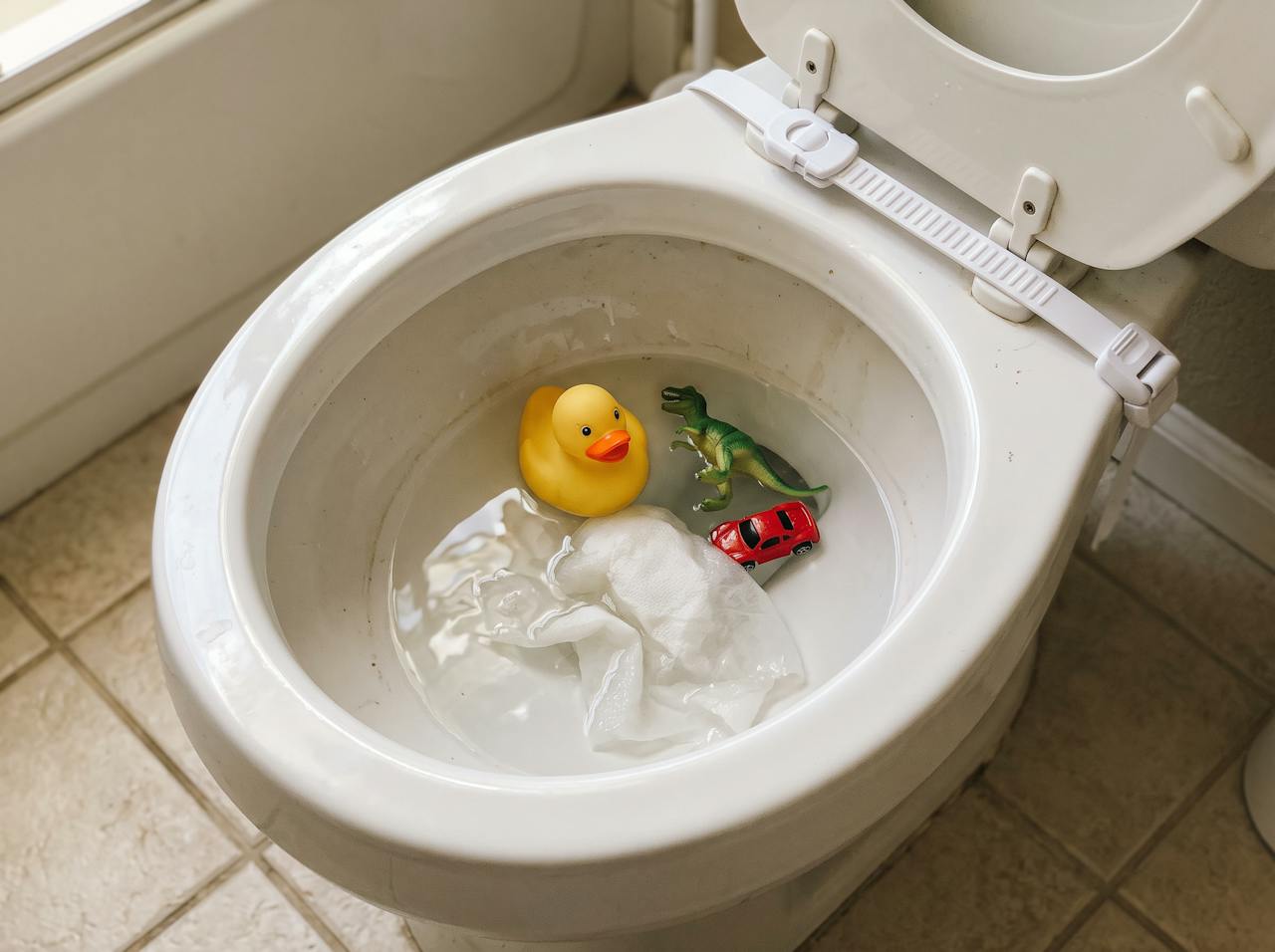 Rubber duck, toy dinosaur, toy car, and a paper towel floating in a toilet bowl with a child safety lock on the lid